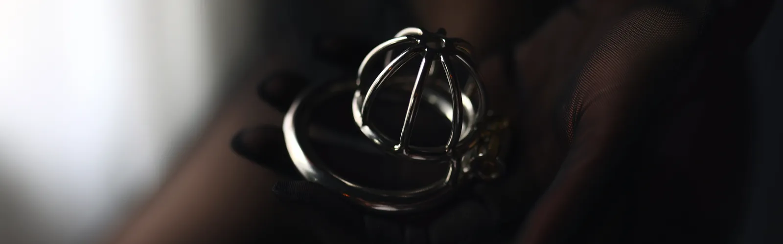 A steel chastity cage resting in a gloved open palm